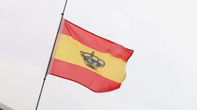 Das Bild zeigt die spanische Flagge auf Halbmast. Die Flagge besteht aus drei horizontalen Streifen – oben und unten rot, mit einem breiteren gelben Streifen in der Mitte, auf dem das spanische Staatswappen abgebildet ist – und steht vor einem bedeckten Himmel, der an das Wetter erinnert, das man während einer Segelreise erleben könnte.