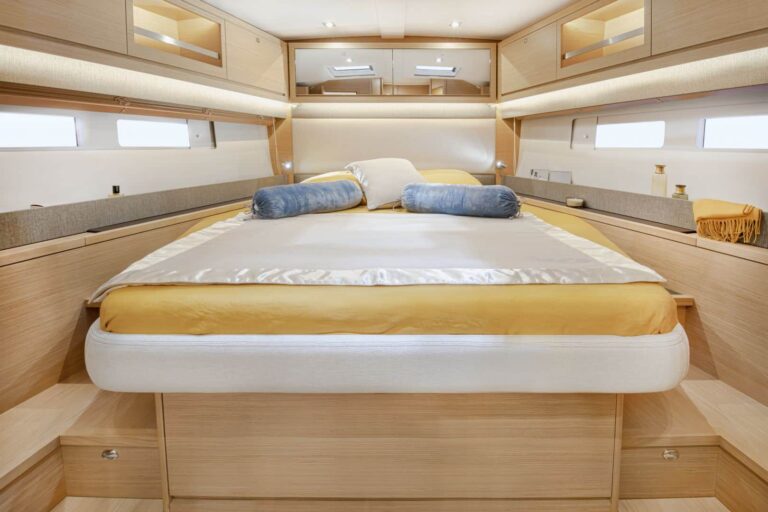 Geräumiges Schlafzimmer in einem Yachtcharter mit modernen Holzmöbeln. Der zentrale Blickfang ist ein großes, ordentlich gemachtes Bett mit gelb-weißer Bettwäsche und blauen Nackenrollen. An den Seiten sind Hängeschränke und kleine Fenster sichtbar, die das helle Ambiente der Kabine verstärken.