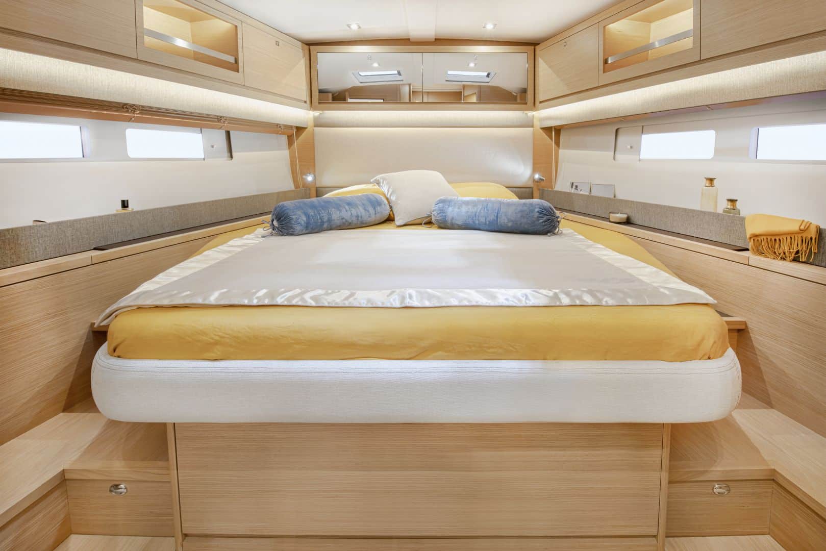 Geräumiges Schlafzimmer in einem Yachtcharter mit modernen Holzmöbeln. Der zentrale Blickfang ist ein großes, ordentlich gemachtes Bett mit gelb-weißer Bettwäsche und blauen Nackenrollen. An den Seiten sind Hängeschränke und kleine Fenster sichtbar, die das helle Ambiente der Kabine verstärken.