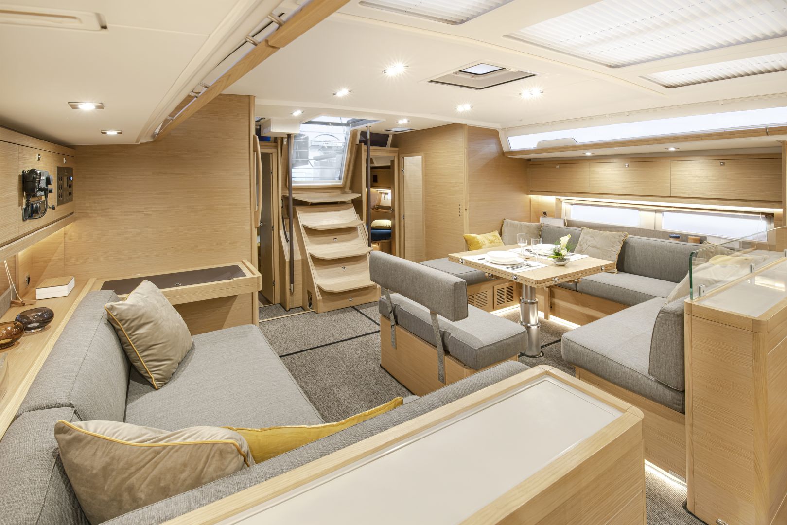 Der Innenraum eines modernen Yachtcharters verfügt über einen geräumigen Wohnbereich mit hellen Holzmöbeln. Es gibt zwei große graue Sofas mit gelben und beigen Kissen, einen hölzernen Esstisch und eine Treppe, die in eine obere Etage führt. Natürliches Licht strömt durch die Fenster darüber herein.