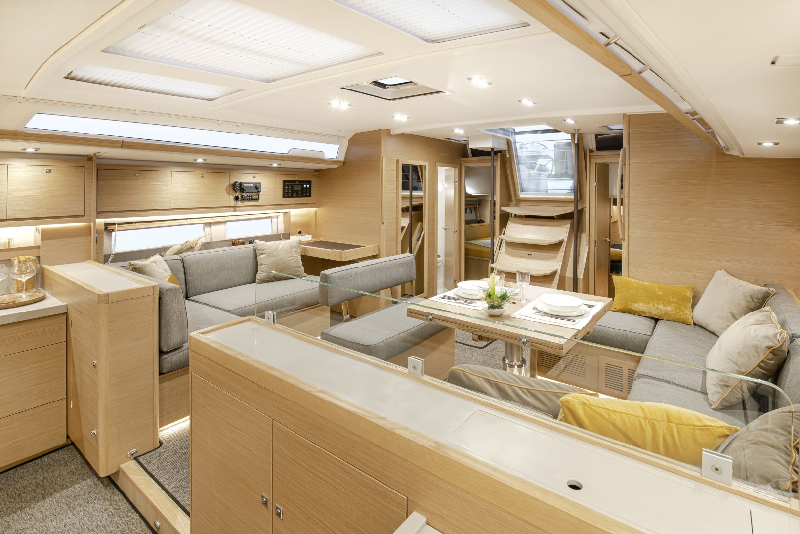 Ein geräumiges, modernes Bootsinterieur mit hellem Holzfinish, ideal für Yachtcharter-Enthusiasten. Der Sitzbereich umfasst grau gepolsterte Bänke mit gelben Kissen. Ein Esstisch ist mit Tellern und Gläsern gedeckt. Dahinter führen Stufen zum Oberdeck. Es gibt reichlich natürliches und künstliches Licht.