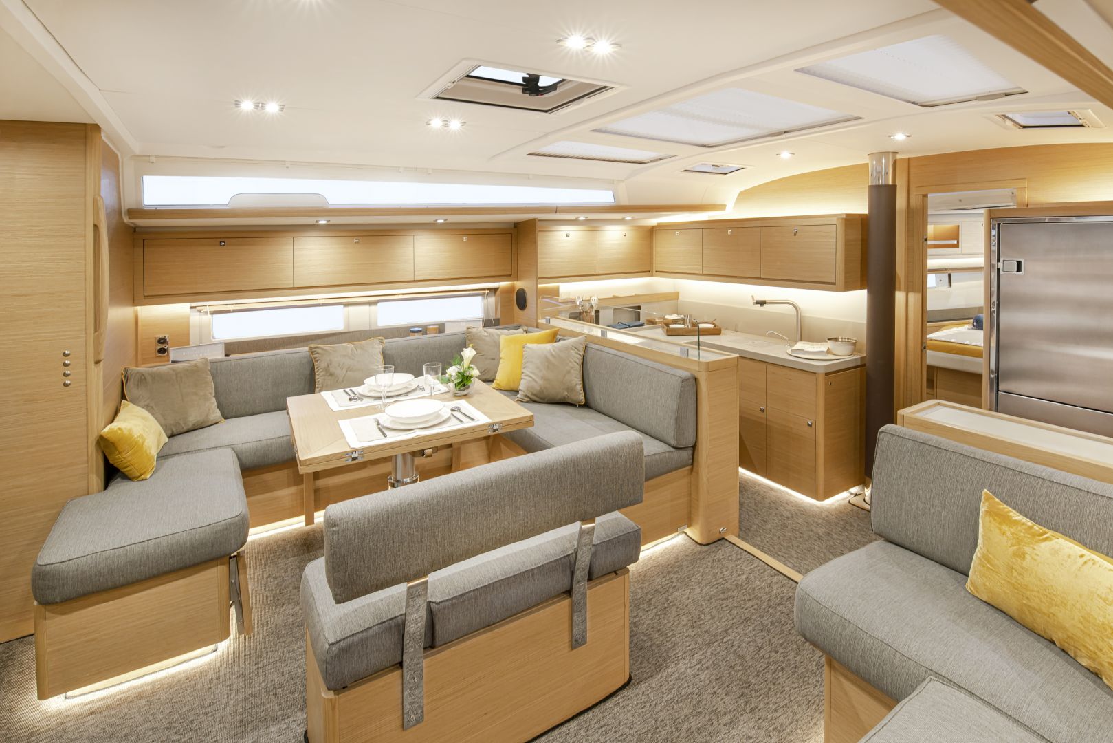 Ein geräumiger Yachtinnenraum steht für Yachtcharter zur Verfügung und bietet einen modernen, offenen Wohn- und Essbereich mit hellen Holzmöbeln und grau gepolsterten Sitzgelegenheiten. Der Bereich umfasst einen Esstisch für zwei Personen, mit einer Küchenzeile im Hintergrund und großen Fenstern, die natürliches Licht hereinlassen.