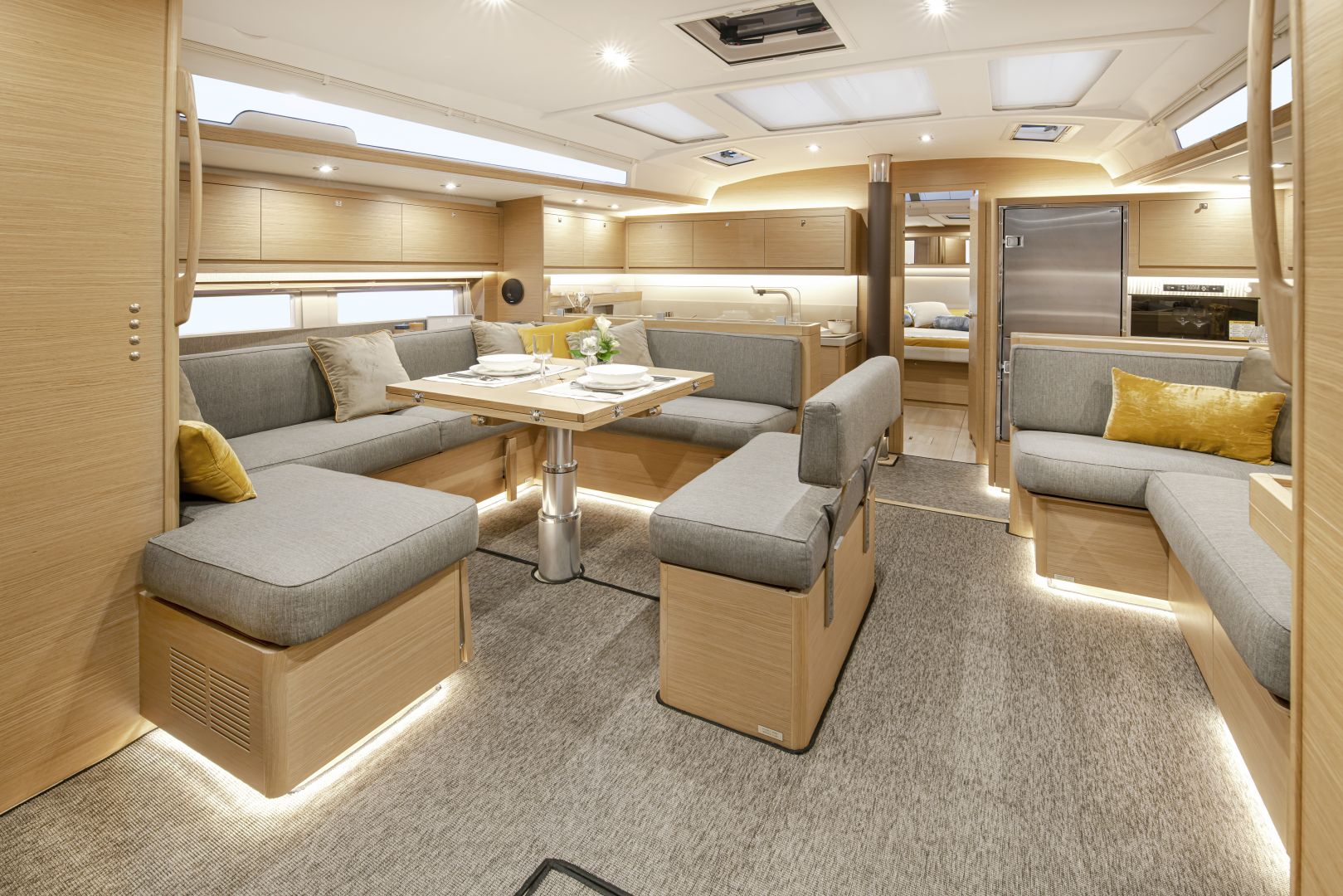 Ein geräumiger, moderner Wohnmobilinnenraum mit hellem Holzfinish. Er verfügt über einen großen, U-förmigen, grau gepolsterten Sitzbereich um einen Esstisch mit Gedecken. Der Raum ist mit Einbauleuchten gut beleuchtet und verfügt im Hintergrund über eine eingebaute Küche, die Yachtcharter-ähnlichen Luxus unterwegs bietet.