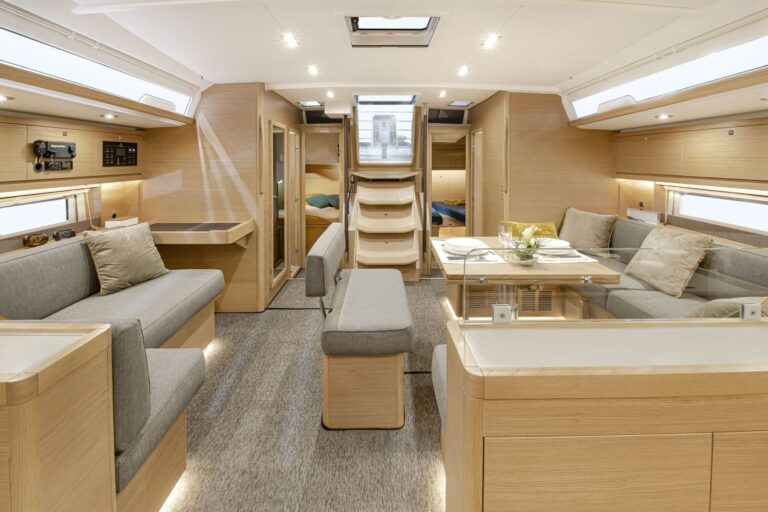 Innenansicht eines modernen Segelboots mit hellen Holzoberflächen und neutralen Farbtönen. Es gibt einen zentralen Sitzbereich mit zwei grauen Sofas, einem kleinen Esstisch und einer Kombüsenküche. Eine Treppe führt zum Schlafbereich. Oberlichter und Fenster sorgen für natürliches Licht – ideal für Yachtcharter-Enthusiasten.
