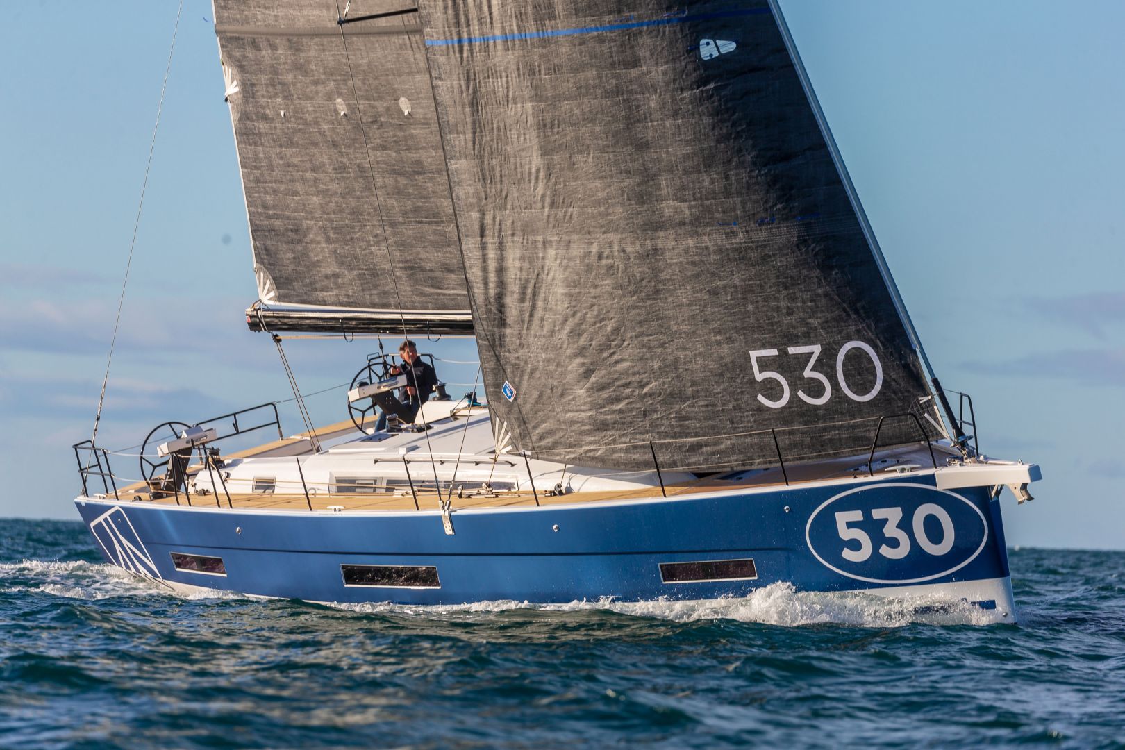 Ein schnittiges blaues Segelboot mit der Nummer 530 auf Rumpf und Segel gleitet sanft über das Wasser. Das für Yachtcharter verfügbare Segelboot hat schwarze Segel und an Deck ist eine einzelne Person zu sehen, die das Schiff manövriert. Das Meer ist ruhig und der Himmel klar.