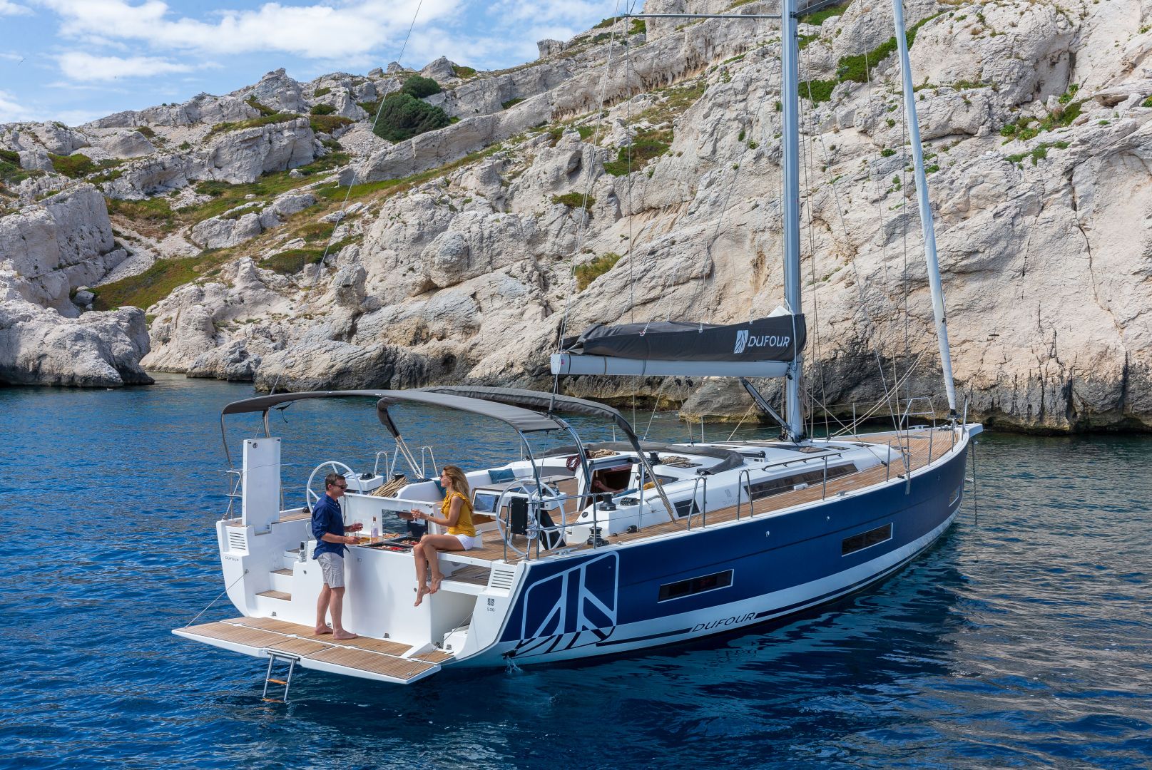 Ein Segelboot ankert in der Nähe von felsigen Klippen und klarem blauen Wasser. Zwei Personen stehen auf dem Deck. Eine Person trägt ein blaues Outfit und die andere ein gelbes. Das Boot, das für Yachtcharter verfügbar ist, weist moderne Designmerkmale und sichtbare Ausstattung auf. Die Sonne scheint und schafft eine ruhige Atmosphäre.
