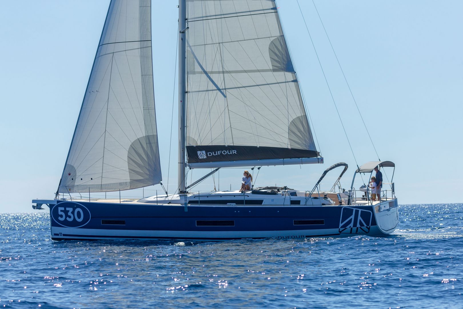 Ein Segelboot mit der Nummer 530 an der Seite segelt auf klarem, blauem Wasser. Das Boot hat zwei große Segel und einen marineblauen Rumpf mit weißen Verzierungen. Auf dem Deck sind zwei Personen zu sehen, die ihren Yachtcharter genießen, und der Himmel darüber ist klar und hell. Auf dem Segel ist ein Logo „DUFOUR“ zu sehen.