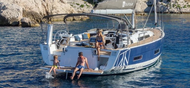 Eine dreiköpfige Familie entspannt sich auf einer gecharterten Yacht im klaren blauen Wasser in der Nähe felsiger Klippen. Das Kind und der erwachsene Mann tauchen ihre Füße hinten im Boot ins Wasser, während eine erwachsene Frau im Badeanzug über ihnen lümmelt. Die Yacht ist blau und weiß.