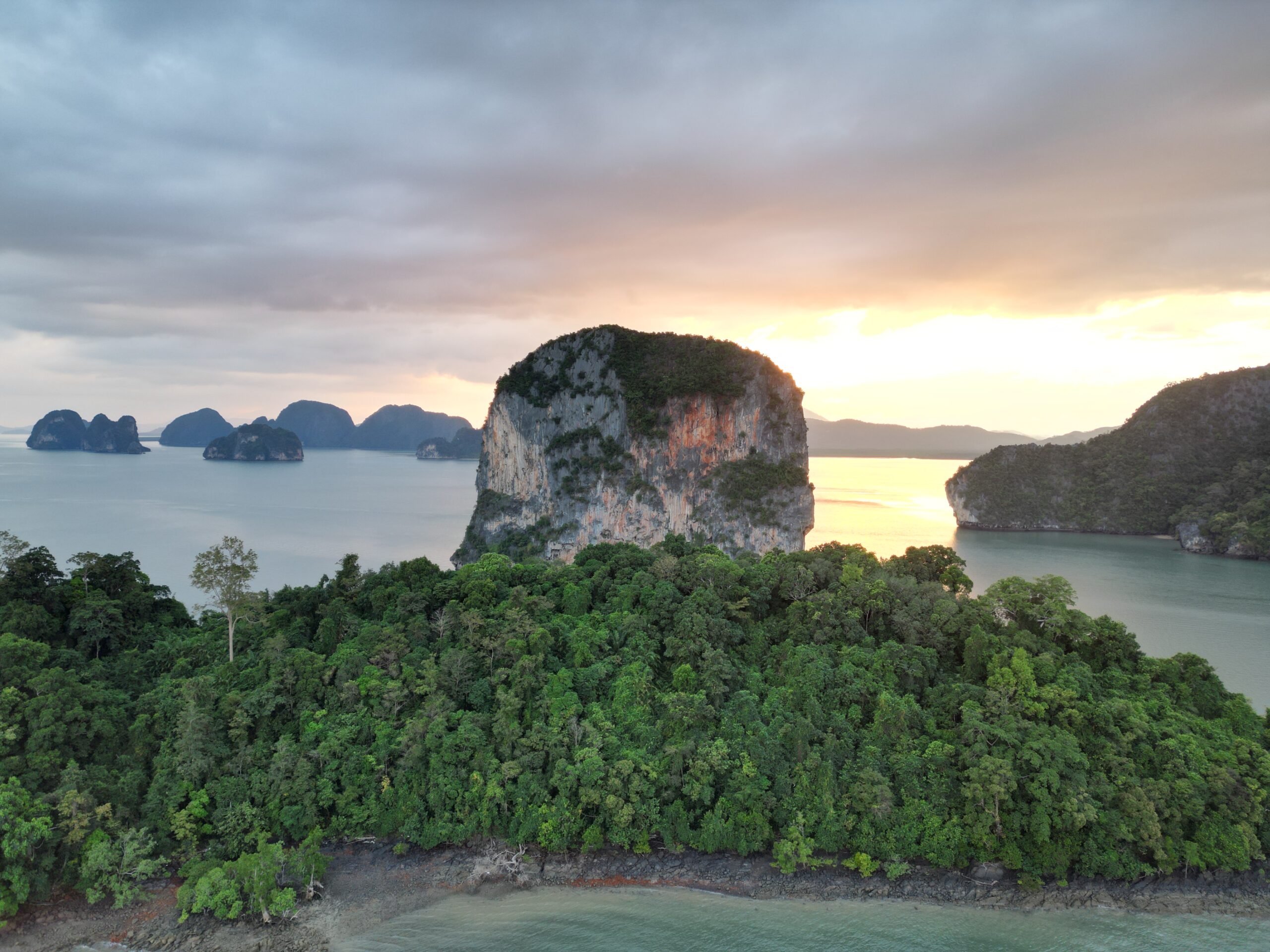 Eine Luftaufnahme einer üppigen, grünen Insel mit steilen Klippen, umgeben von ruhigem, türkisfarbenem Wasser bei Sonnenuntergang. Ideal für einen Wasserurlaub, der Horizont zeigt weitere kleinere Inseln unter einem dramatischen, bewölkten Himmel mit Flecken goldenen Sonnenlichts.