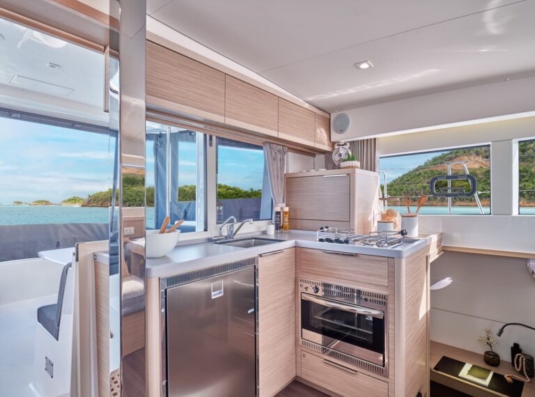 Eine moderne Bootsküche auf einem Youngline-Katamaran mit hellen Holzschränken, einer Edelstahlspüle, einem Herd mit Backofen und einer Arbeitsplatte mit Utensilien. Ein Fenster bietet einen Blick auf üppige grüne Hügel und blaues Wasser. Helle und luftige Atmosphäre mit natürlichem und künstlichem Licht – eine perfekte Kulisse für Ihr Segeltörn-Abenteuer.