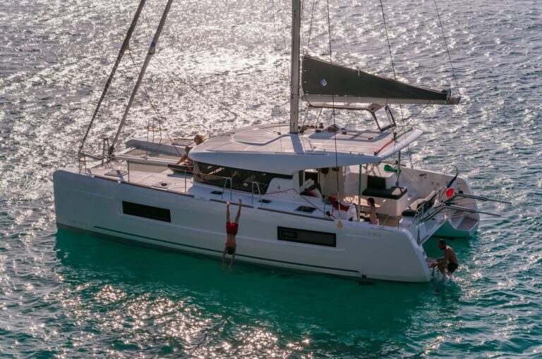 Ein weißer Youngline-Katamaran mit mehreren Personen an Bord liegt im klaren, türkisfarbenen Wasser vor Anker. Eine Person hängt an der Seite, während eine andere in der Nähe schwimmt. Die Sonne spiegelt sich im Wasser und erzeugt einen glitzernden Effekt. Der Katamaran hat ein Segel und mehrere sichtbare Annehmlichkeiten, perfekt für einen Segeltörn.