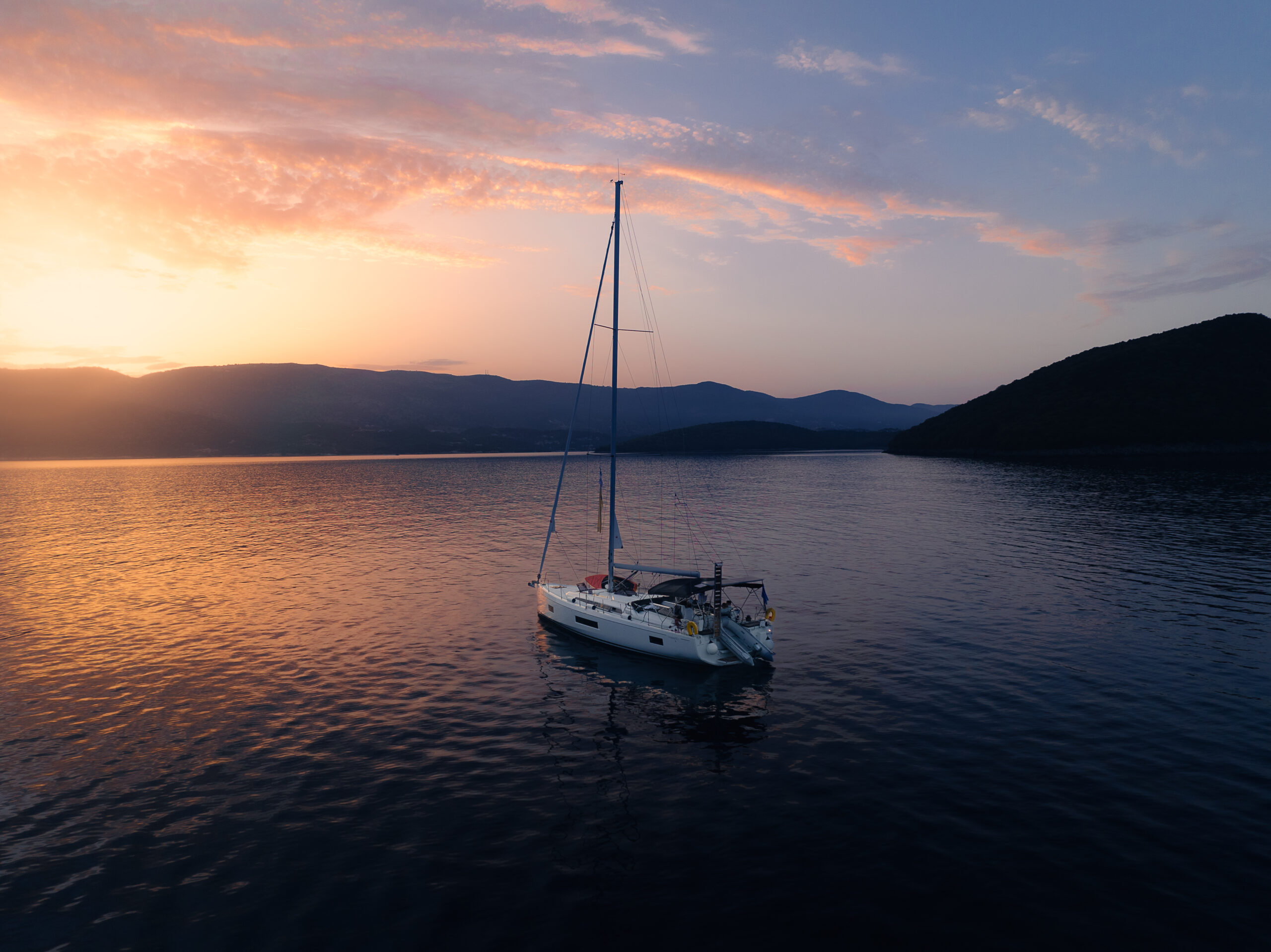 Ein Segelboot, das bei Sonnenuntergang auf ruhigem Wasser vor Anker liegt, perfekt für einen Segelurlaub. Der Himmel ist mit einer Mischung aus Orange-, Lila- und Rosatönen geschmückt, während die Berge in der Ferne eine ruhige Kulisse bilden. Die friedliche Szene strahlt Ruhe und natürliche Schönheit aus.
