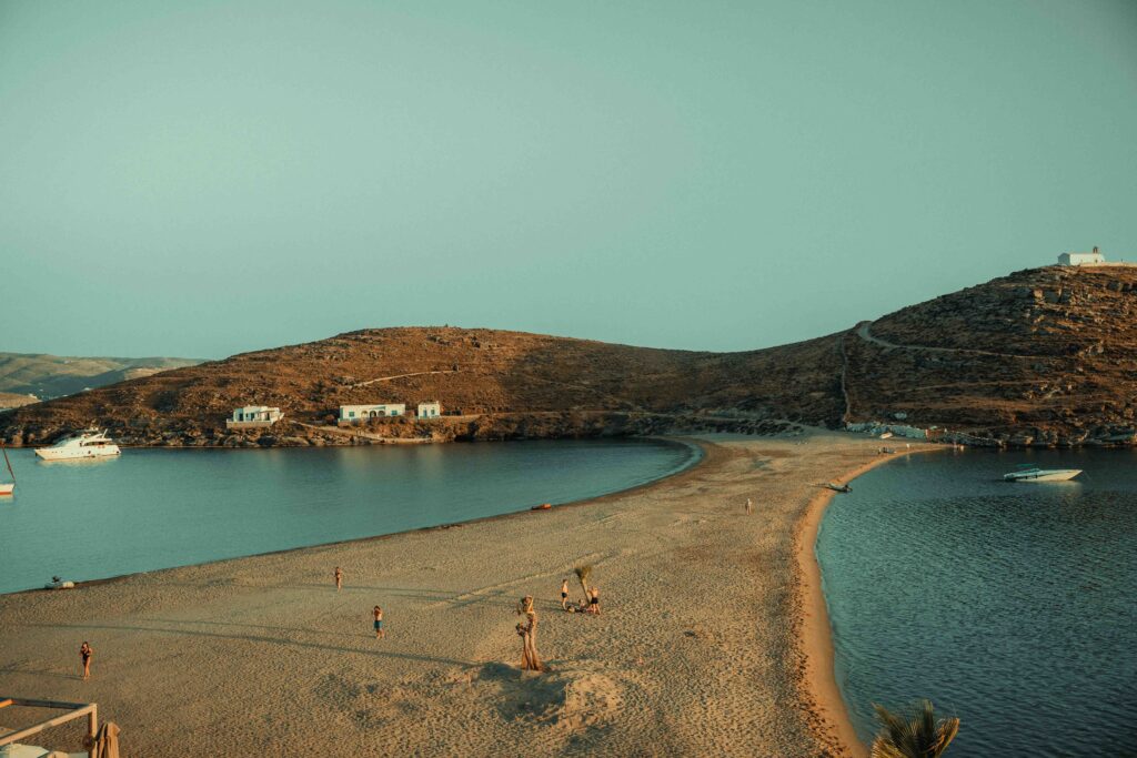 Between two bodies of water stretches a calm, narrow strip of beach where a few people walk and relax. In the background, hills with sparse vegetation rise under a clear sky, reminiscent of the tranquil Cyclades islands, with a few small white buildings dotting the shoreline.