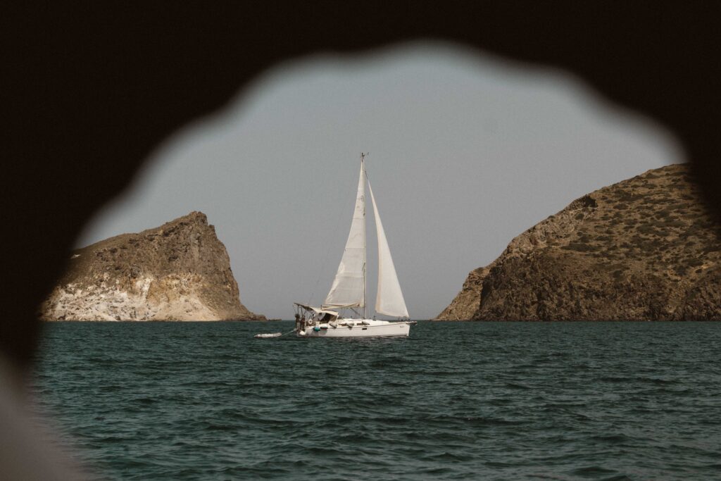A white sailboat glides over calm seas, framed by rocky cliffs on both sides. The scene, reminiscent of a sailing adventure, is viewed through a keyhole-shaped opening with the sailboat centered. The sky is clear, and the water is a deep blue-green.