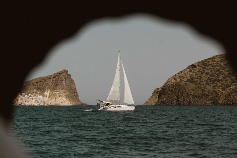 Ein weißes Segelboot gleitet auf einem ruhigen Meer, eingerahmt von felsigen Klippen auf beiden Seiten. Das Bild, das an ein Segelabenteuer erinnert, wird durch eine schlüssellochartige Öffnung betrachtet, mit dem Segelboot in der Mitte. Der Himmel ist klar und das Wasser ist tief blaugrün.