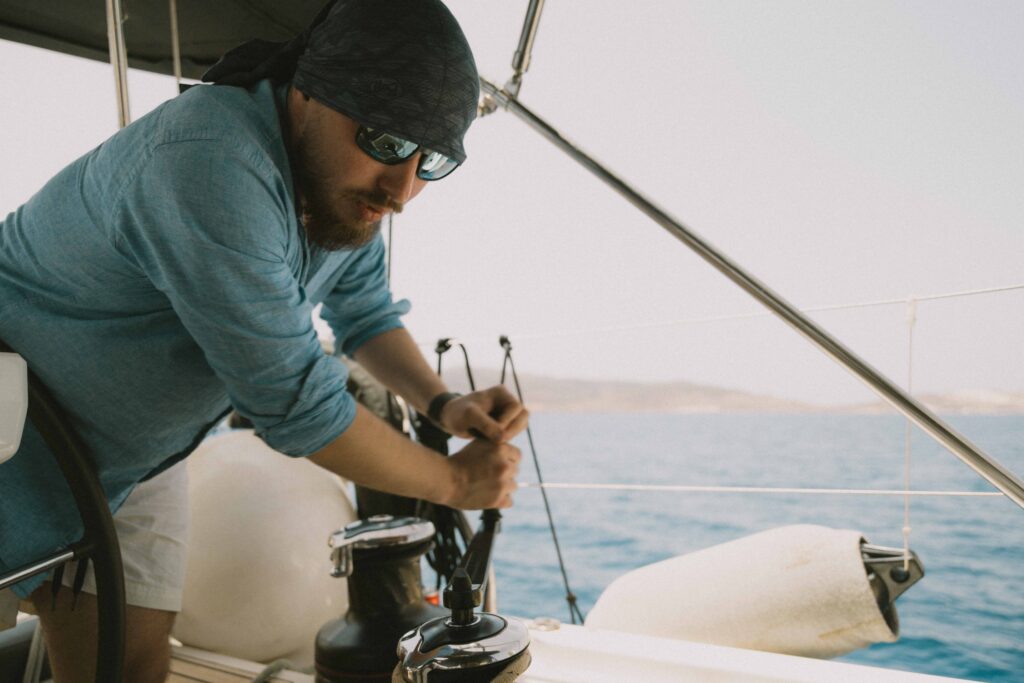 A man wearing a headscarf and sunglasses operates the sails of a sailboat on a sunny day. He’s dressed in a light blue long-sleeve shirt and white shorts, enjoying the sailing while calm blue waters sparkle behind him and the coastline is visible in the distance.
