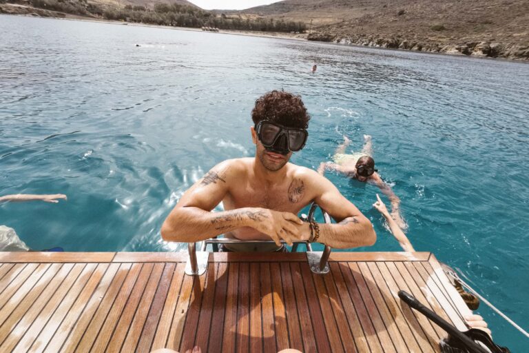 Eine Person mit Schwimmbrille steht auf einer Holzplattform am Rande des türkisfarbenen Wassers, im Hintergrund sind andere Schwimmer und vorbeigleitende Segelboote zu sehen. Die Plattform erstreckt sich bis ins Meer, umgeben von felsigen Hügeln unter einem teilweise bewölkten Himmel.