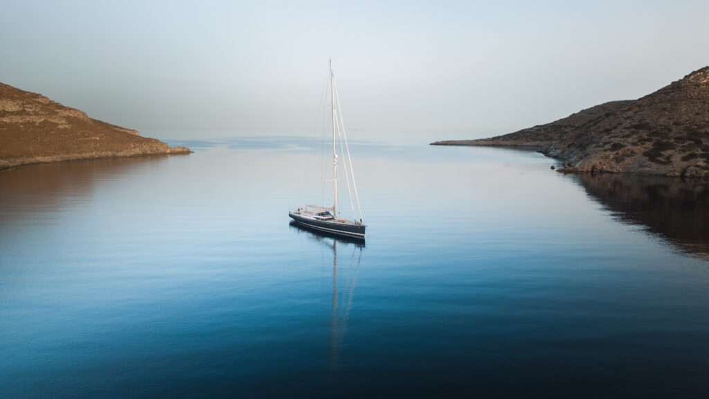 A solitary sailboat floats on calm, reflective blue water between two rugged, brown islands. The sky over the Cyclades is clear and bright, adding to the peaceful atmosphere of the scene.