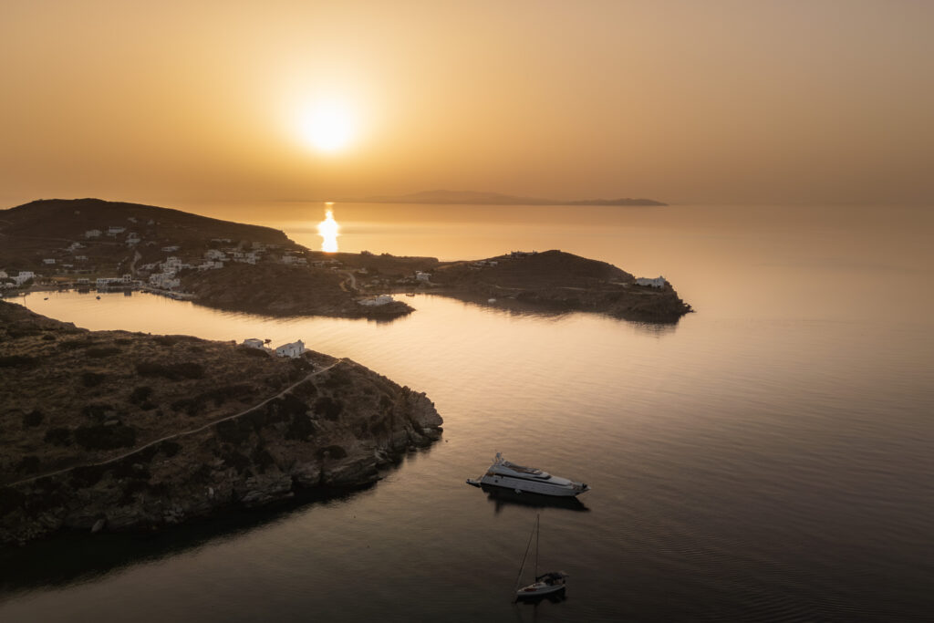 A cheerful sunset over calm seas, with two boats anchored near rocky shores. The reflected sun creates a golden path on the water, perfect for evening sailing, while the coastline features a few white buildings and rugged hills in the background. The sky is painted in warm shades of orange.