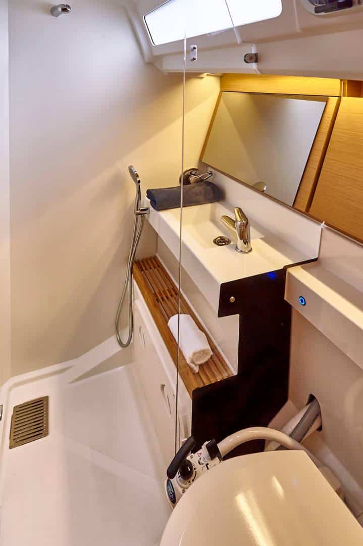 Ein kompaktes, modernes Bootsbadezimmer mit weißen und hölzernen Oberflächen. Es verfügt über ein Waschbecken mit einem Spiegel darüber, einen Duschkopf, ein kleines Handtuch auf einem Holzregal und eine Toilette im Vordergrund. Der Raum ist gut beleuchtet, mit natürlichem Licht, das durch ein Fenster hereinfällt – ideal für jedes Yachtcharter-Erlebnis.