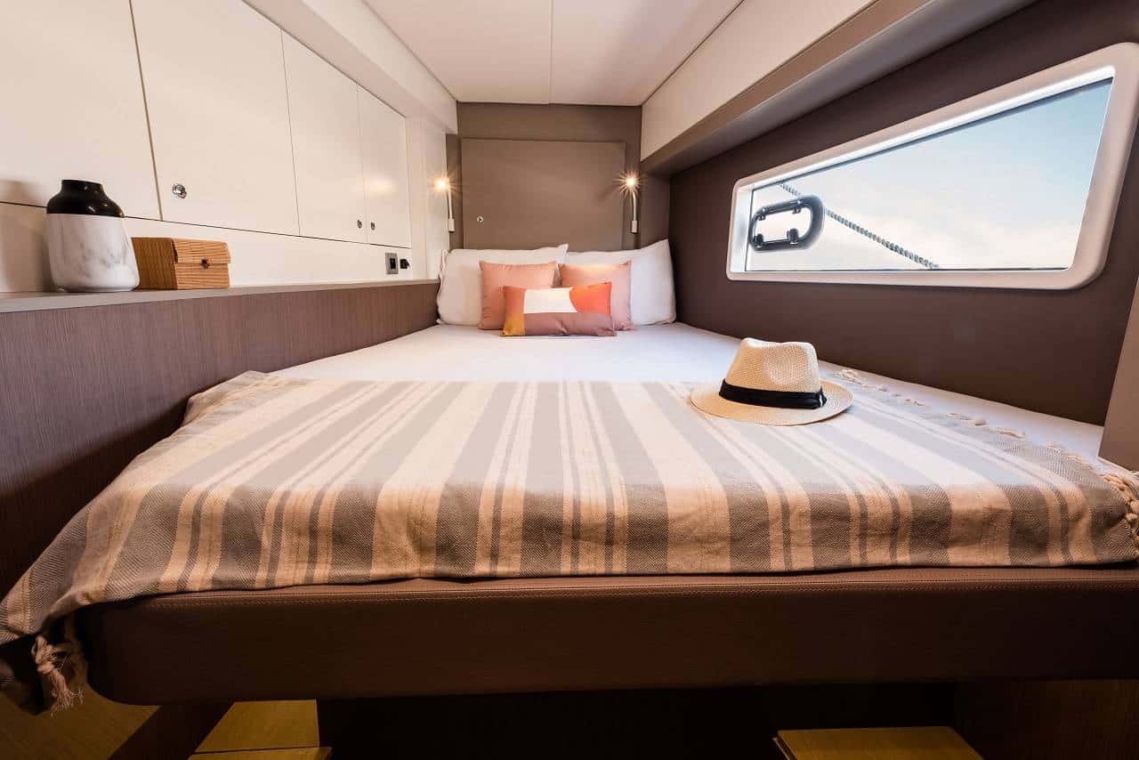 Ein ordentlich gemachtes Bett mit gestreifter Bettwäsche in einer Kabine mit Holz- und Weißakzenten. Auf dem Bett liegen ein Hut und ein buntes Kissen. Die Kabine verfügt über Einbauschränke, ein Fenster mit Blick auf den Youngline Katamaran Segeltörn und gezielte Beleuchtung am Kopfende des Bettes.