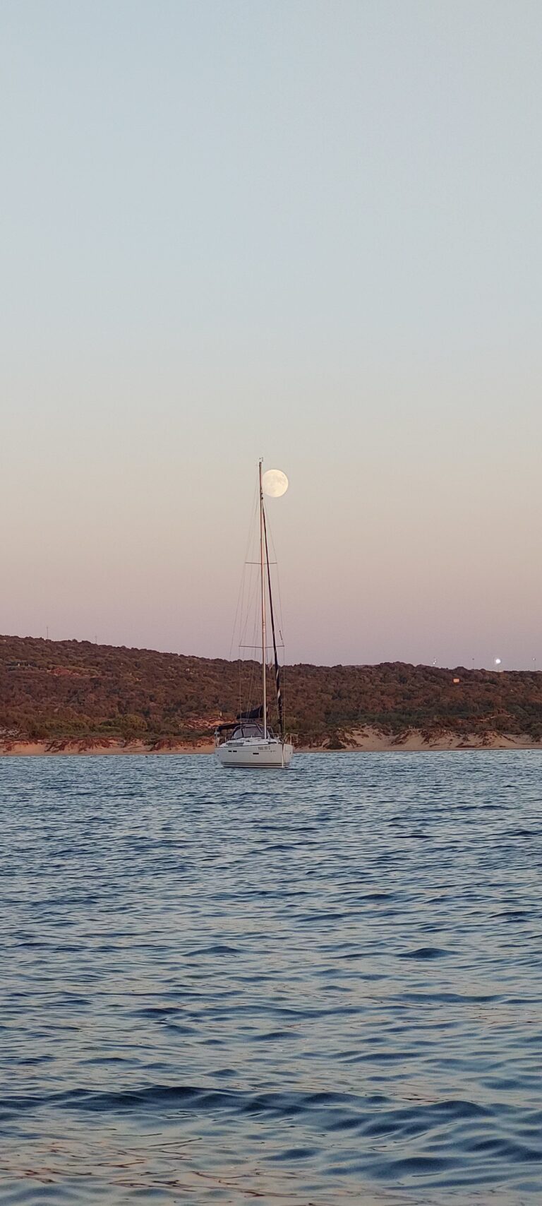 Ein Segelboot vor Anker in ruhigen Gewässern mit felsigen Hügeln im Hintergrund, das die ruhige Atmosphäre eines Segelurlaubs auf Sardinien einfängt. Der Vollmond steht tief am Himmel und erweckt den Eindruck, als säße er auf dem Mast des Segelboots. Der Himmel ist in sanften Farbtönen gehalten, die entweder Sonnenaufgang oder Sonnenuntergang anzeigen.