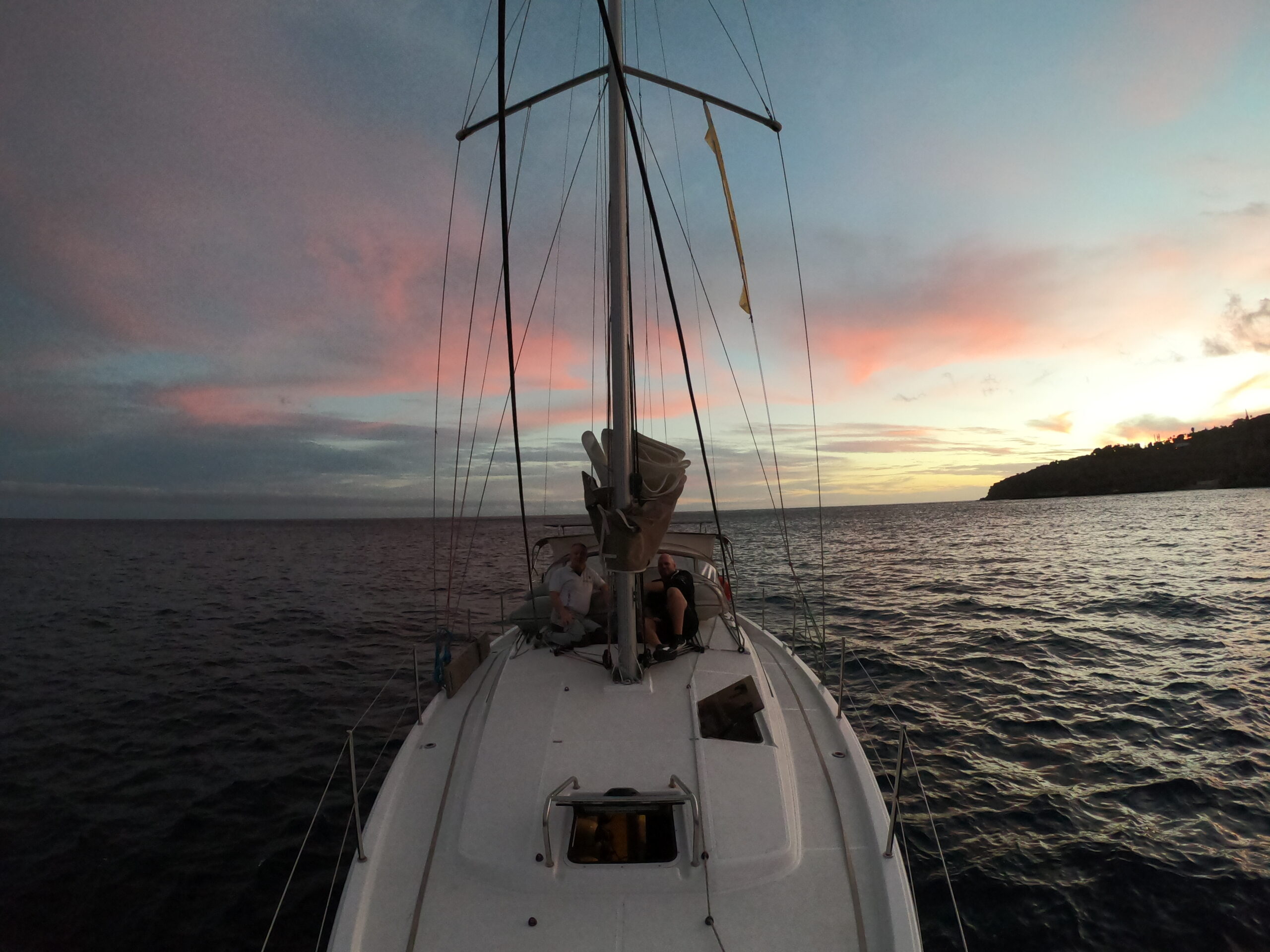A person sits on the deck of a sailboat at sea, gazing at a calm sunset painted in shades of pink, blue, and violet. During the Atlantic crossing from Gibraltar to Tenerife, the boat heads toward a distant coastline on the horizon. The calm sea and colorful sky create a peaceful atmosphere.  