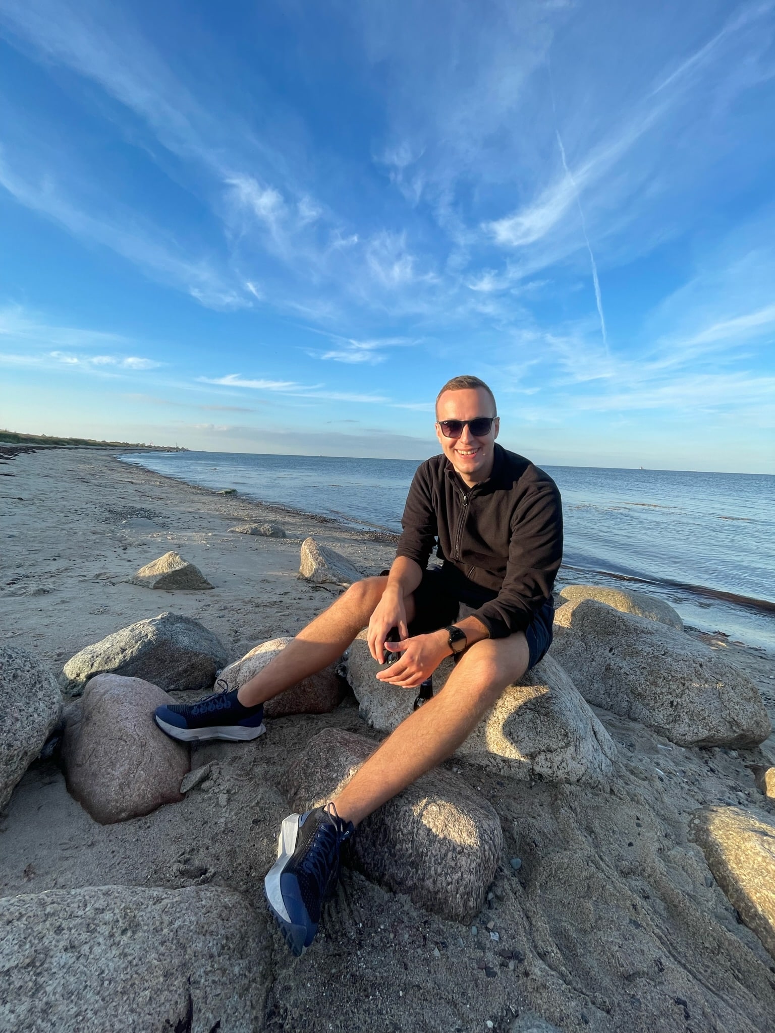 Eine Person mit Sonnenbrille, schwarzem Langarmshirt und Shorts sitzt an einem sonnigen Tag auf einem Felsen an einem ruhigen Strand. Der Himmel ist blau mit dünnen Wolken und im Hintergrund ist ruhiges Wasser am Ufer zu sehen. Die Person lächelt und schaut in die Kamera, während sie ihre Youngline Segelreise genießt.