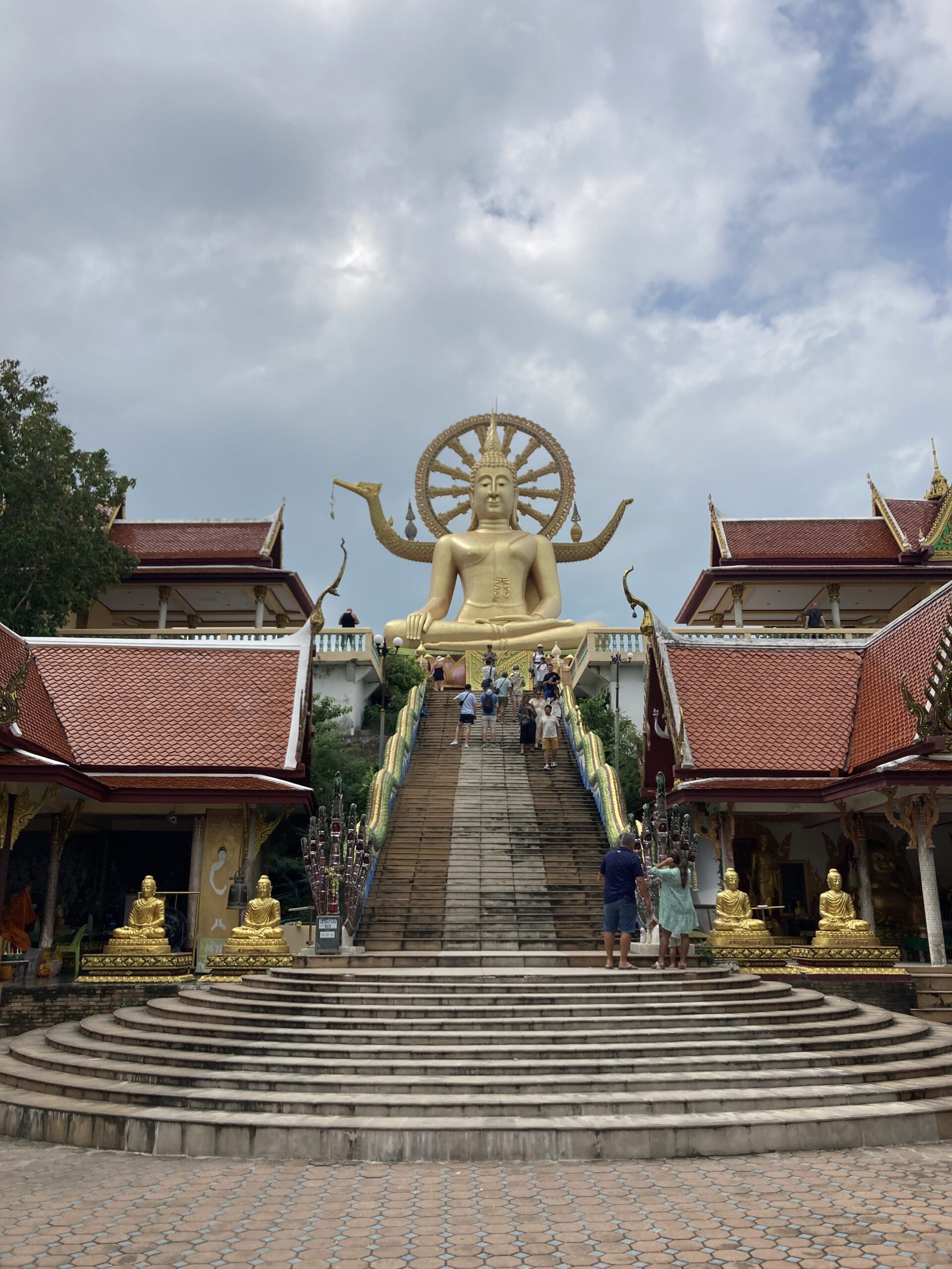 A large golden Buddha statue stands at the top of a wide staircase, flanked by ornate buildings with red roofs. Several people, perhaps telling stories of their last sailing trip to Greece, climb the stairs. The sky above is cloudy with patches of blue.