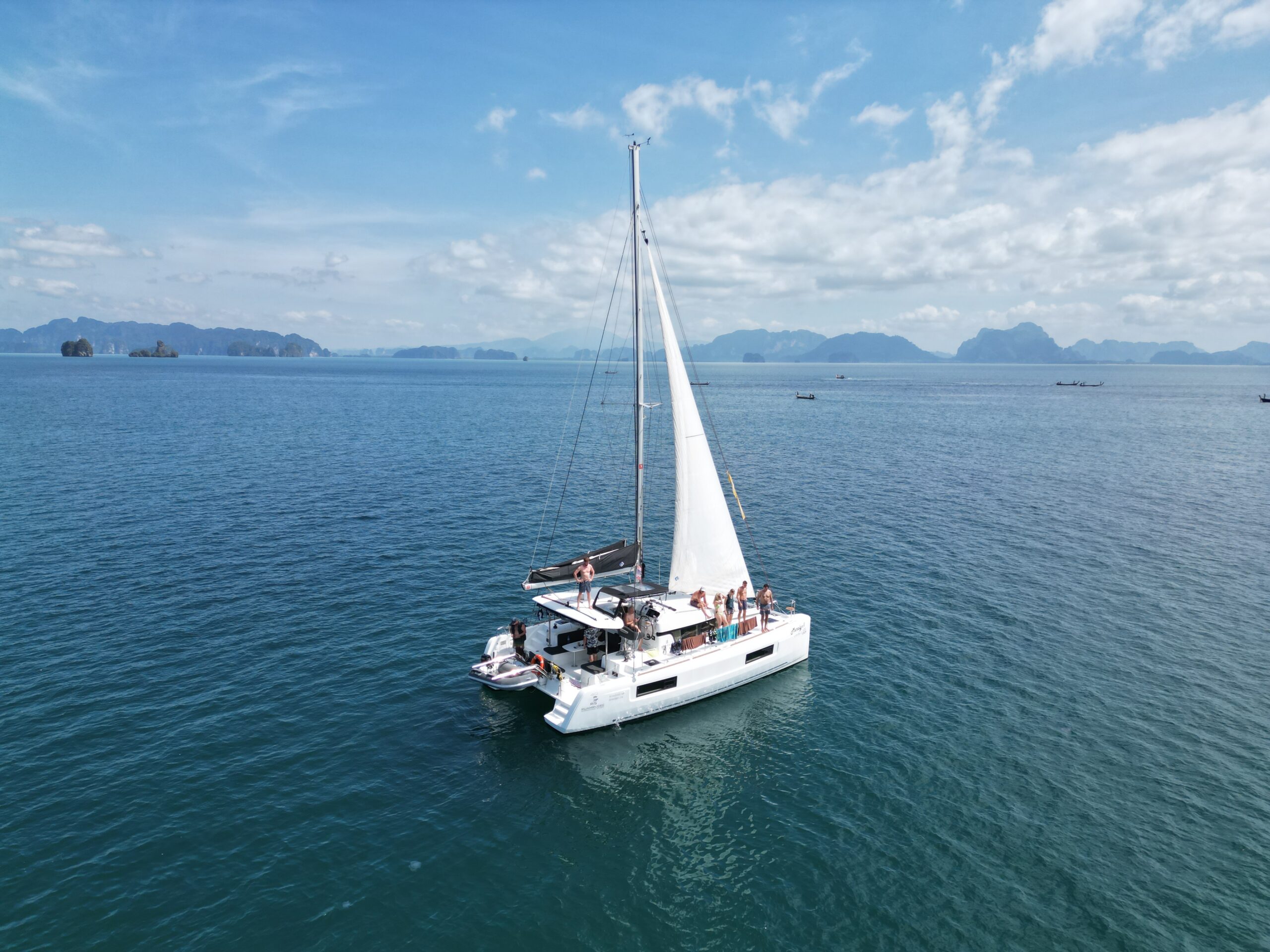 Ein weißes Segelboot mit einer Gruppe von Menschen an Bord kreuzt in den ruhigen blauen Gewässern vor Phuket, Thailand, mit einer Kulisse aus fernen Inseln unter einem teilweise bewölkten Himmel. Das Segel ist vollständig ausgefahren und die Szene zeigt ein ruhiges und malerisches Segeln.