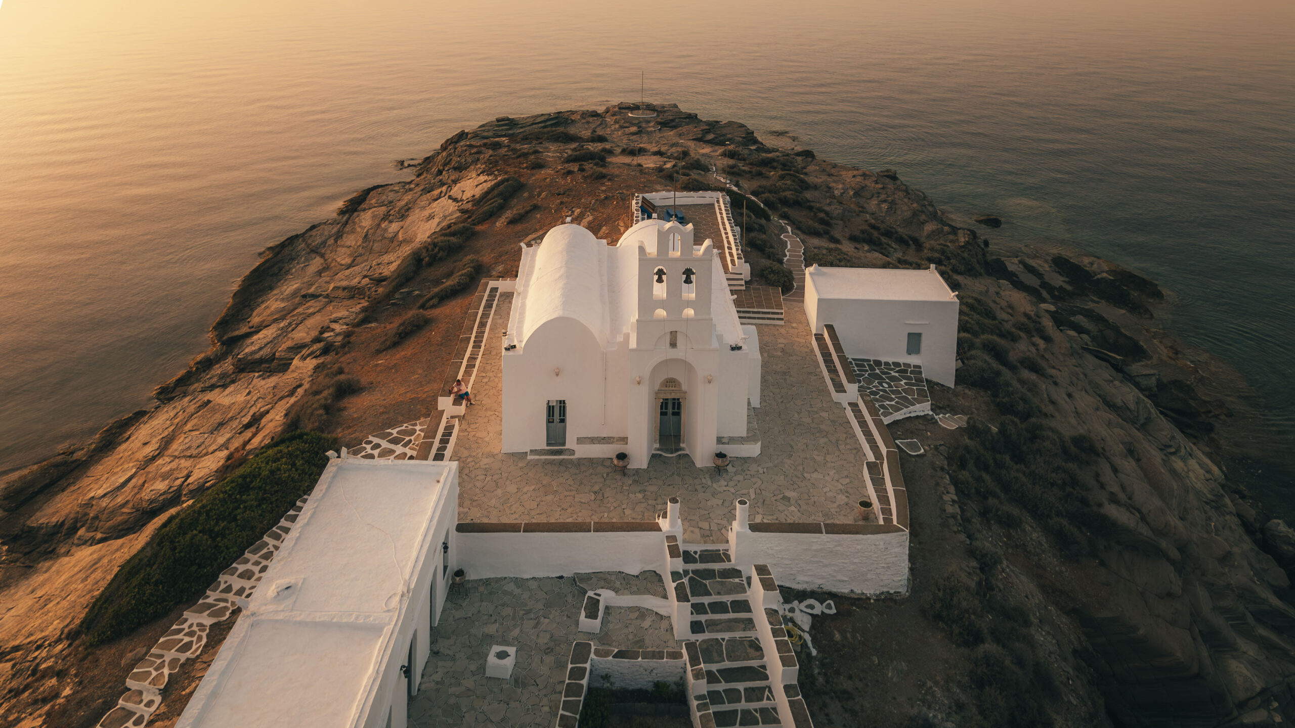 Aerial view of a white chapel with arched windows and a small bell tower near the cliff edge overlooking a calm sea at sunset. The rocky landscape and stone paths around the chapel add to the peaceful, picturesque atmosphere - perfect for anyone embarking on a sailing trip to Greece.