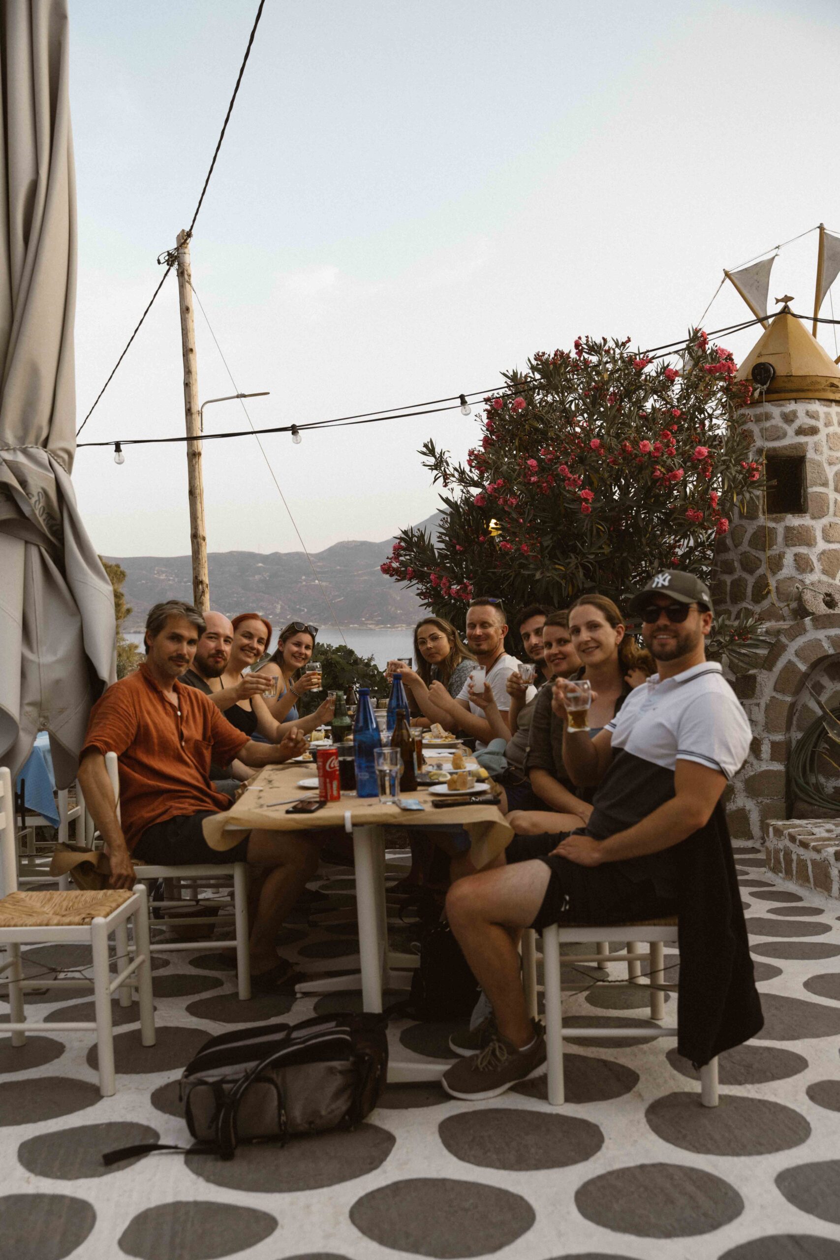 A group of people sit around a long outdoor table, smiling and clinking glasses. The backdrop consists of a stone floor, a windmill, flowering bushes and fairy lights overhead. In the background are rolling hills under a clear sky, reminiscent of a peaceful sailing trip to Greece.