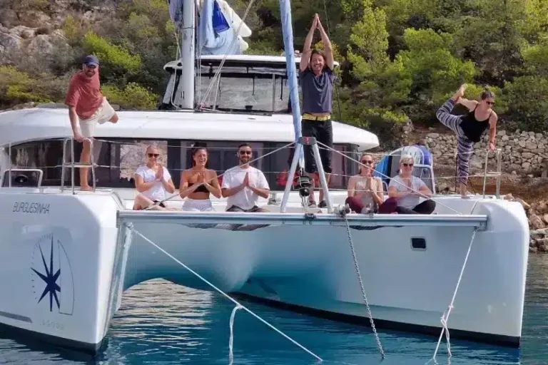 Eine Gruppe von Menschen vergnügt sich auf einer Segelyacht, die in der Nähe einer felsigen Küste vor Anker liegt. Einige sitzen, andere stehen oder posieren, alle wirken fröhlich unter dem sonnigen Himmel.