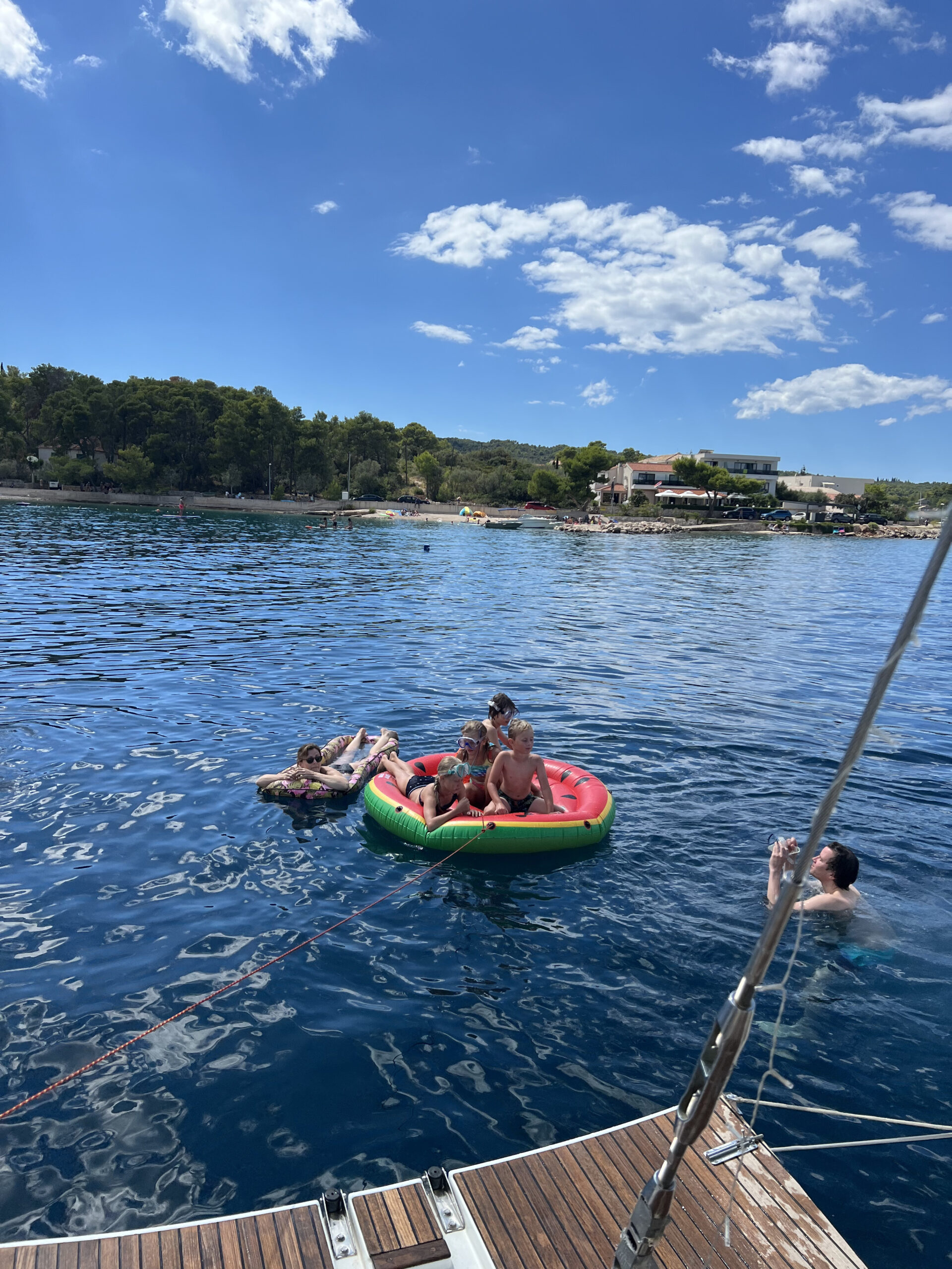 Vier Menschen genießen einen sonnigen Tag auf dem Wasser. Drei von ihnen sitzen auf einem großen, aufblasbaren rot-grünen Schwimmkörper, während eine Person in der Nähe schwimmt. Im Hintergrund ist die Uferlandschaft mit Bäumen, Gebäuden und ein paar Menschen zu sehen, was den Charme des familienfreundlichen Segeltörns im Mittelmeer hervorruft.