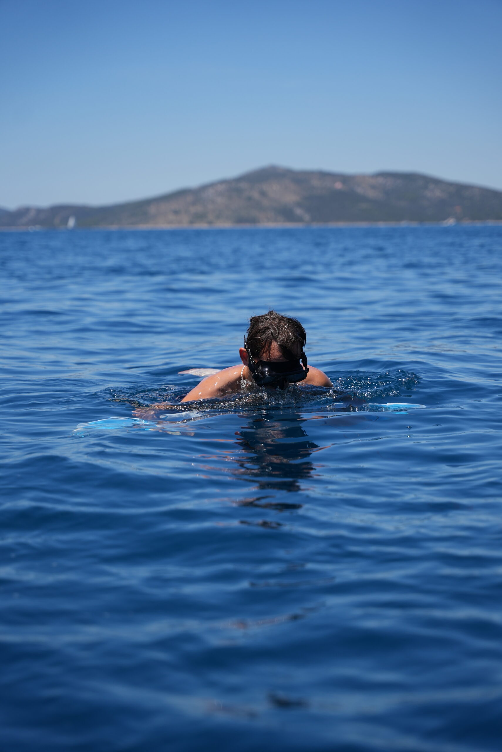 A person swims in a vast, clear blue sea, with their head above the water. They are wearing swim goggles and seem to be looking down. In the distance, a mountainous island under a clear blue sky hints at an adventurous sailing experience in Croatia.  