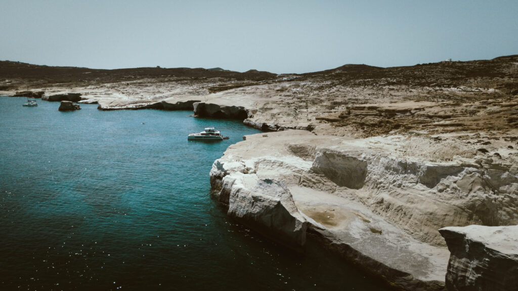 A peaceful coastal scene with clear turquoise waters and rugged white cliffs of the Cyclades. A few boats float near the shore, with a larger one anchored close to the rocks. The landscape in the background is dry, with minimal vegetation, under a clear blue sky.