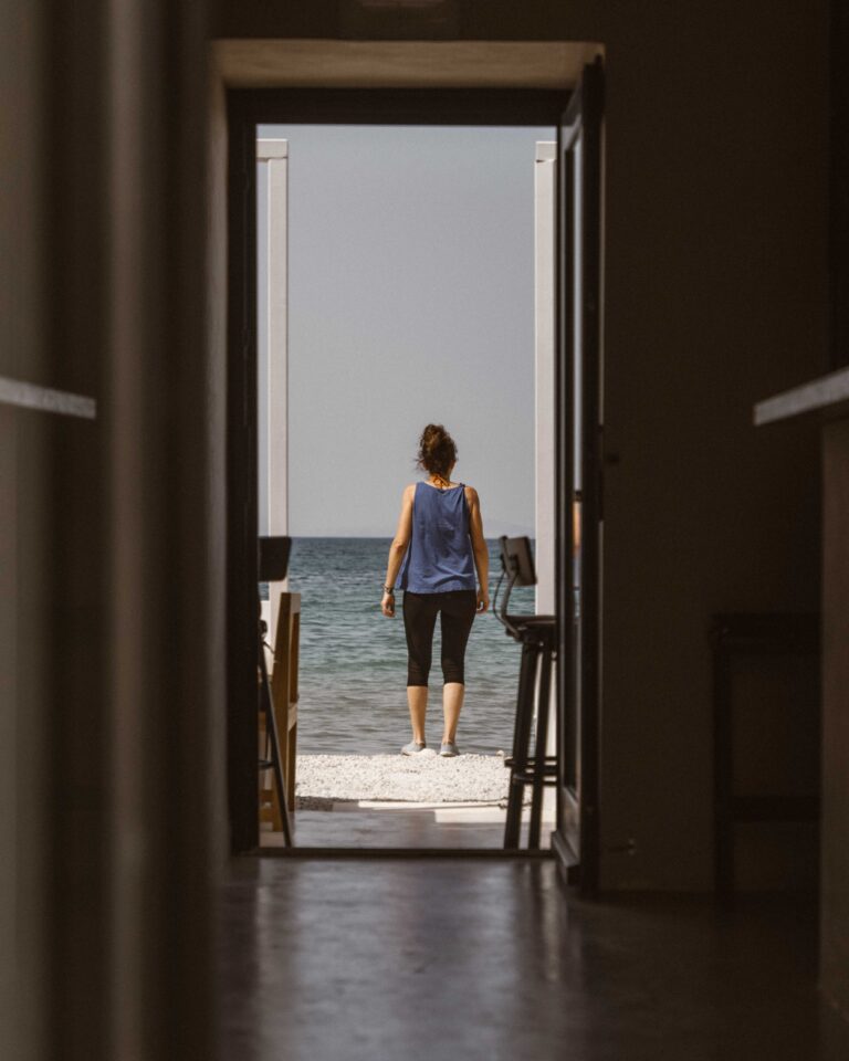 Eine Person in einem blauen Tanktop und schwarzen Leggings geht an einem Kiesstrand zum Meer. Sie ist durch eine offene Tür in einem schwach beleuchteten Flur zu sehen. Auf beiden Seiten des Flurs sind Möbel zu sehen, die auf eine Segeltour hindeuten. Die Szene ist hell und sonnig.