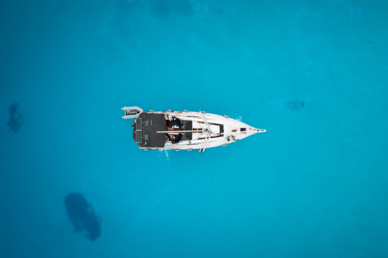 Luftaufnahme eines einzelnen weißen Segelboots, das in klarem, leuchtend blauem Wasser schwimmt. Das Boot befindet sich in der Bildmitte, umgeben von der unendlichen Weite des Meeres, und zeigt seine Ruhe und Abgeschiedenheit – eine heitere Segeltour auf ruhigem Wasser.