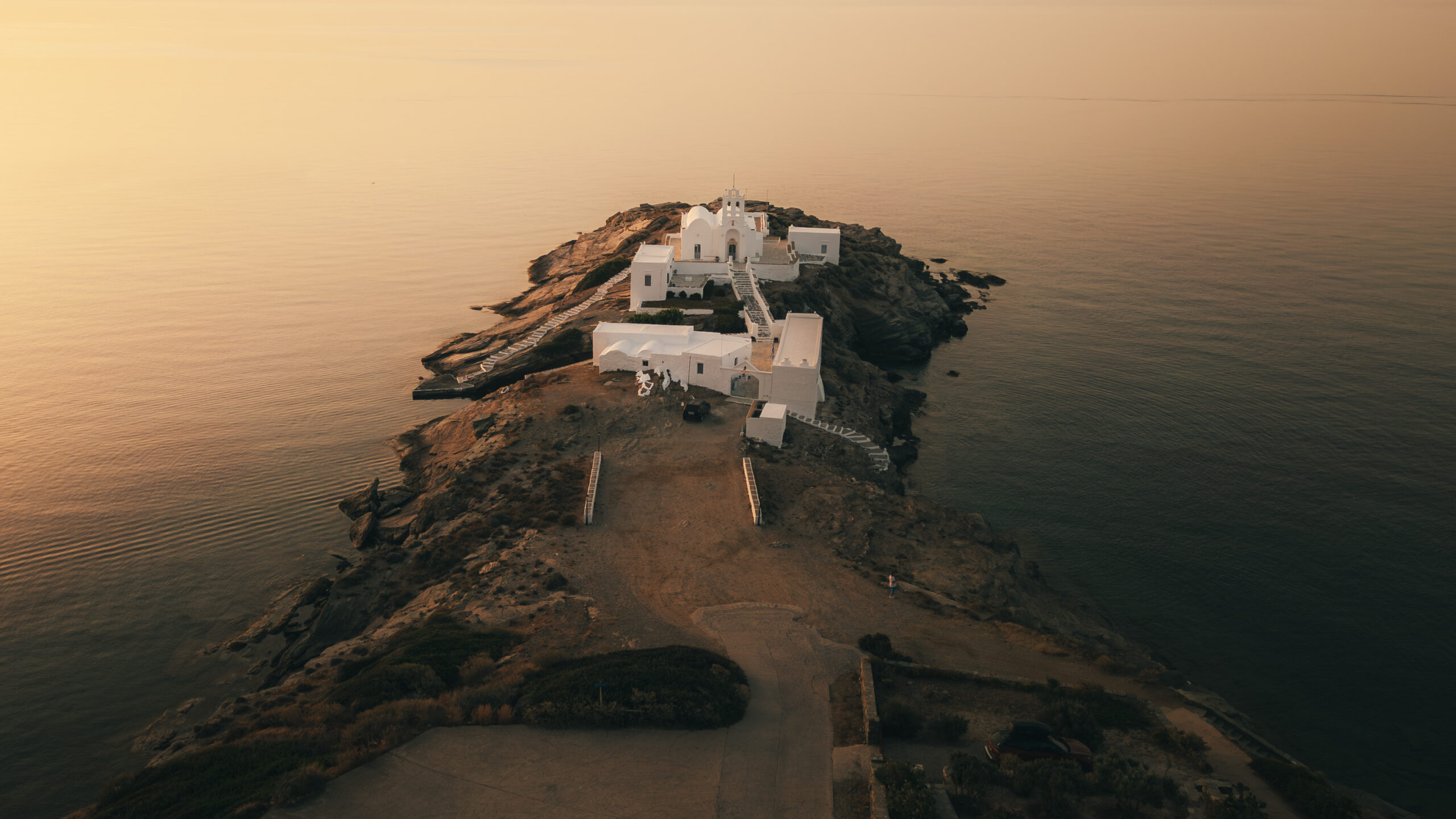 Luftaufnahme einer Halbinsel bei Sonnenuntergang. Weiße Gebäude drängen sich an der Spitze der Landform, umgeben von ruhigem, reflektierendem Wasser. Die Szene ist in ein warmes, goldenes Licht getaucht. Spärliche Vegetation und felsiges Gelände führen zu den Gebäuden – ein idyllischer Anblick für diejenigen auf einer Segeltour.