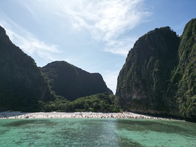 Eine wunderschöne tropische Strandszene mit klarem türkisfarbenem Wasser im Vordergrund. Der Strand ist voller Menschen und wird auf beiden Seiten von üppigen, grünen Klippen gesäumt, was an ein Segelreise-Abenteuer in Griechenland unter einem strahlend blauen Himmel mit dünnen Wolken erinnert.