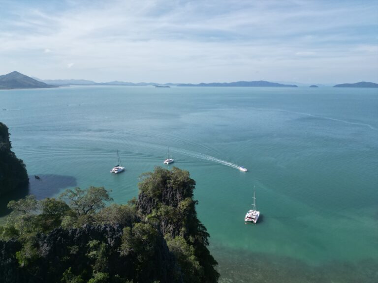 Vier Segelboote liegen verstreut auf einem weiten, ruhigen blauen Meer, umgeben von felsigen, bewaldeten Inseln. Ein paar dünne Wolken zieren den klaren Himmel und der Horizont ist von fernen Landmassen gesäumt. Die Szene fängt eine heitere Meereslandschaft ein, die an ein ruhiges Segelreise-Abenteuer in Griechenland erinnert.