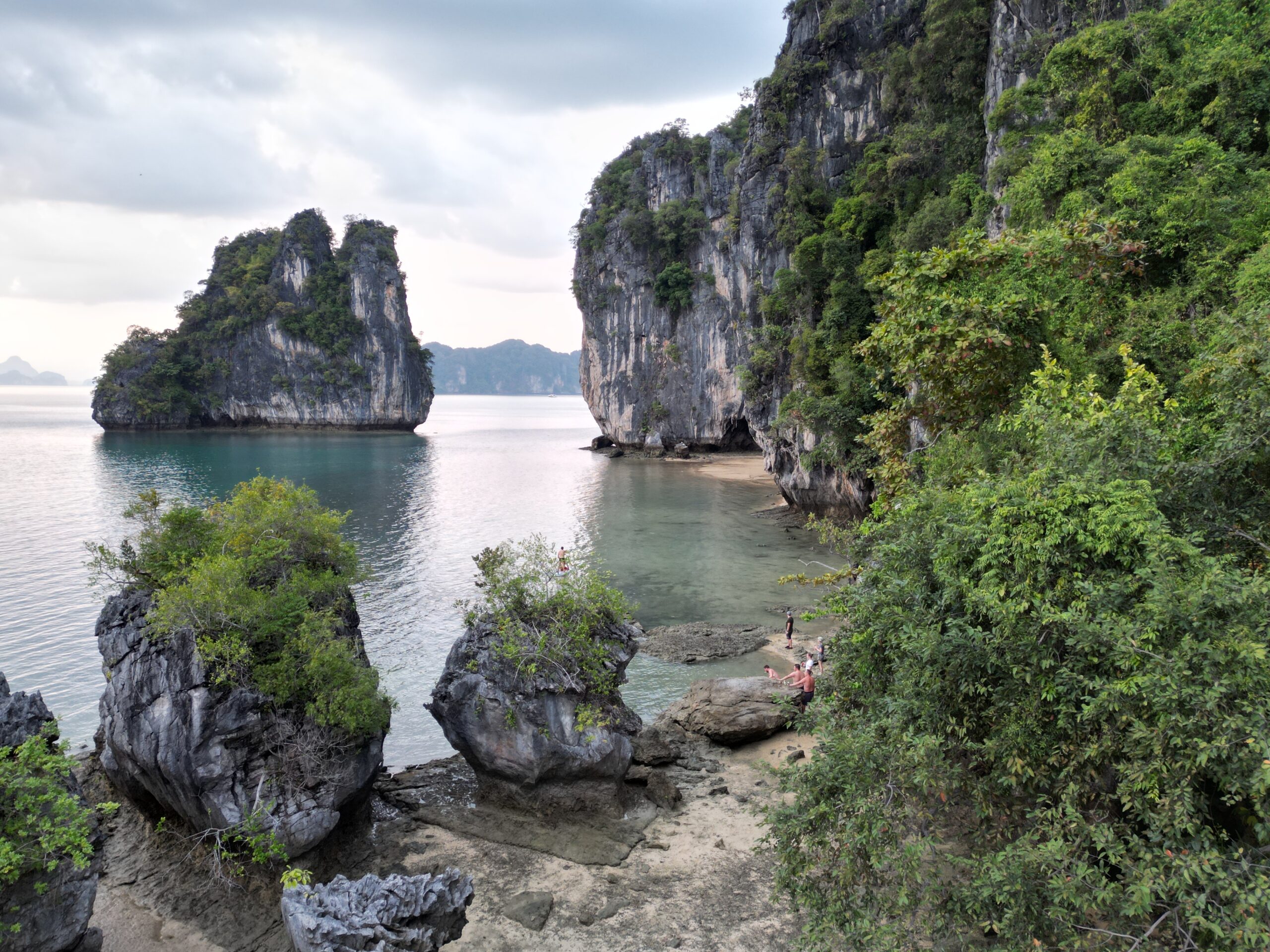 A peaceful coastal scene in Greece with towering limestone cliffs, partially covered in greenery. Clear, calm water surrounds the rocks and reflects the cloudy sky. Small sandy beaches are visible between the cliffs. A sailing trip in Greece shows distant figures sitting on a rock ledge.   