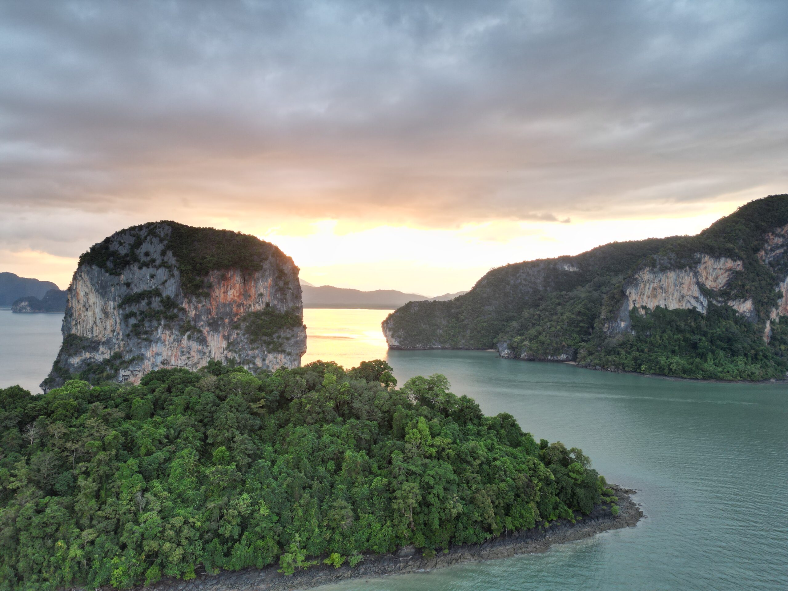 Aerial view of lush green islands, surrounded by calm green water at sunset. The sky is partly cloudy, and the sun casts a golden hue over the landscape. Perfect for a sailing trip to Greece. The scene includes a large, forested rock formation in the foreground and more islands in the background.   