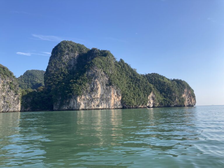 Ein heiterer Blick auf ein ruhiges, grünlich-blaues Meer mit großen, üppigen, grün bewachsenen Kalksteinklippen unter einem klaren blauen Himmel, der an eine Segelreise nach Griechenland erinnert. Die Sonne beleuchtet Teile der Klippen, hebt ihre Texturen hervor und erzeugt Spiegelungen im Wasser.