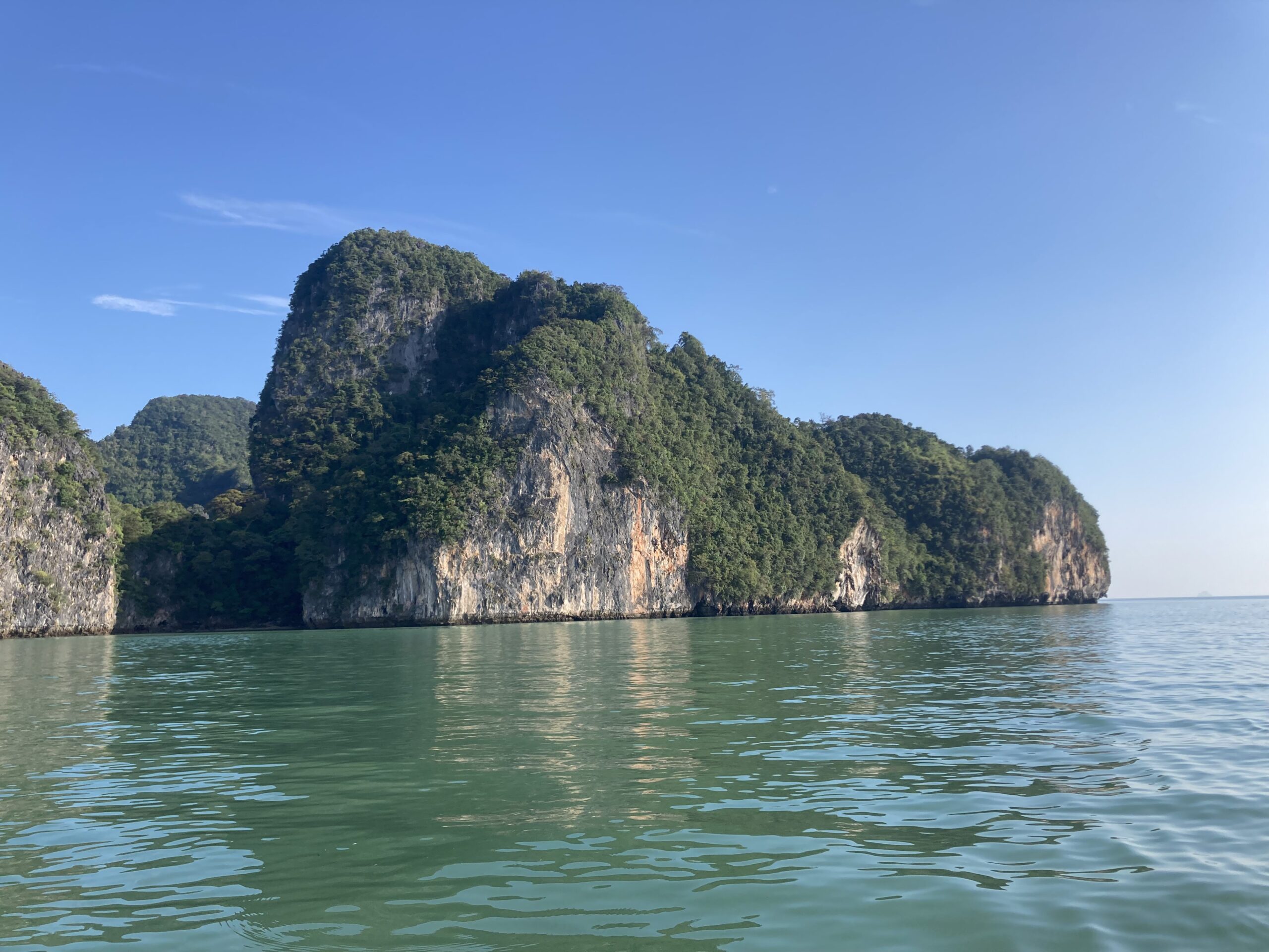 Ein heiterer Blick auf ein ruhiges, grünlich-blaues Meer mit großen, üppigen, grün bewachsenen Kalksteinklippen unter einem klaren blauen Himmel, der an eine Segelreise nach Griechenland erinnert. Die Sonne beleuchtet Teile der Klippen, hebt ihre Texturen hervor und erzeugt Spiegelungen im Wasser.