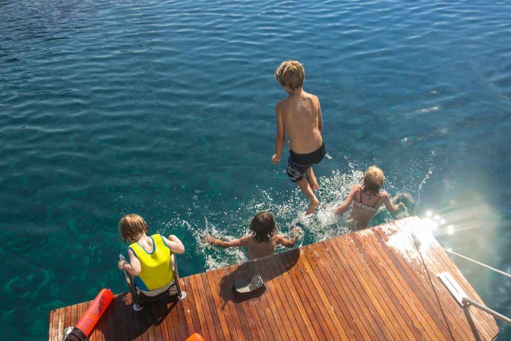 Four children are enjoying the water: one climbs a wooden dock ladder, while the other three are either jumping in or splashing in the clear blue water on a sunny day. The scene evokes family-friendly sailing trips in the Mediterranean and represents fun and adventure.