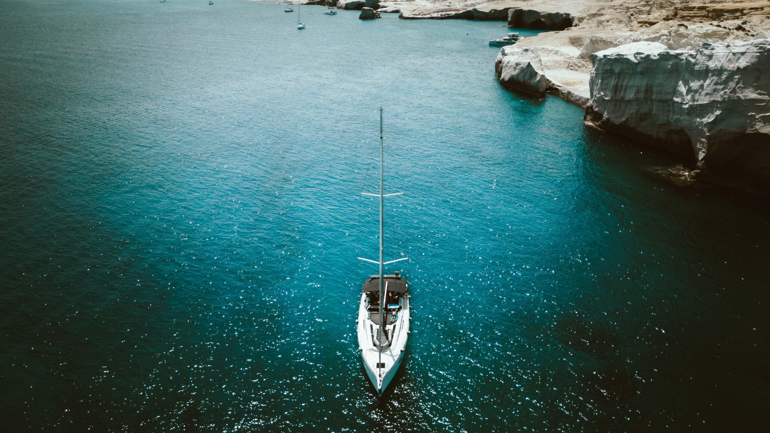 A white sailing boat floats on a clear, deep blue sea near a rocky coast with white cliffs. The water glistens in the bright sunlight and other smaller boats can be seen in the distance. It is an ideal backdrop for skipper training in the Mediterranean and makes the scenery both tranquil and picturesque.  