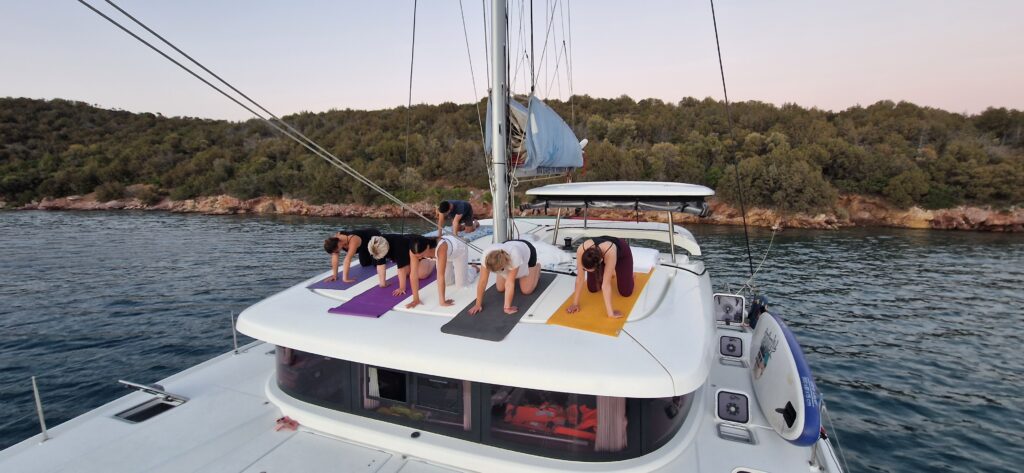 Eine Gruppe von Menschen praktiziert Yoga auf dem Deck eines Segelboots, das in der Nähe einer bewaldeten Küste vor Anker liegt. Der Himmel ist klar und von der untergehenden Sonne in einen sanften rosa Farbton getaucht. Die Teilnehmer stehen auf Yogamatten, balancieren auf Händen und Füßen und genießen die Ruhe nach einem Tag Skippertraining im Mittelmeer.