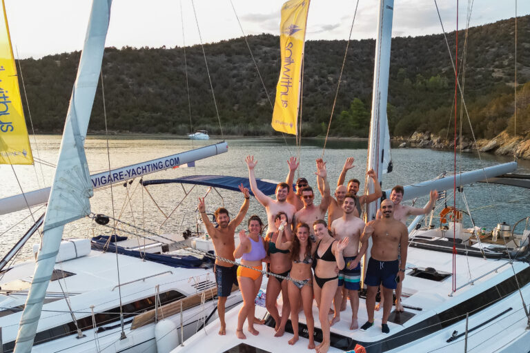 Eine Gruppe von Menschen steht lächelnd und winkend auf dem Deck einer weißen Yacht. Sie tragen Badebekleidung und scheinen einen sonnigen Tag während ihres Segelurlaubs auf Sardinien zu genießen. Neben ihnen ist eine weitere Yacht zu sehen, im Hintergrund sind malerische Hügel und Bäume zu sehen.