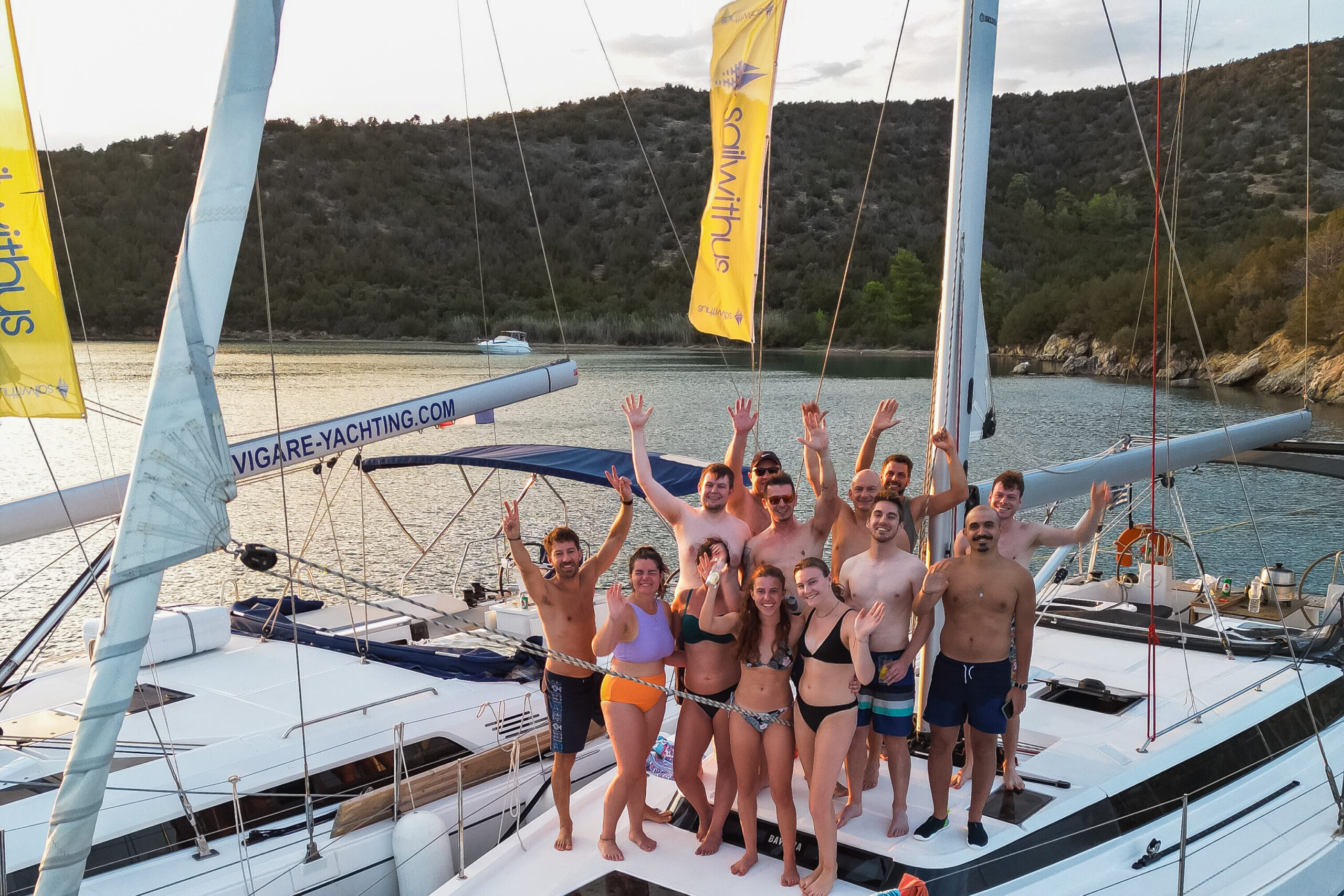 Eine Gruppe von Menschen steht lächelnd und winkend auf dem Deck einer weißen Yacht. Sie tragen Badebekleidung und scheinen einen sonnigen Tag während ihres Segelurlaubs auf Sardinien zu genießen. Neben ihnen ist eine weitere Yacht zu sehen, im Hintergrund sind malerische Hügel und Bäume zu sehen. Aktivurlaub